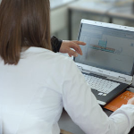 Training BiMOTiON Eine Frau in weißer Bluse sitzt vor einem Laptop. Von links zeigt eine Hand einer anderen Person auf den Bildschirm.